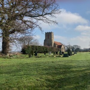 A photograph of a country church, St Peters Corpusty, Norfolk. Set in a green churchyard with blue sky and trees.