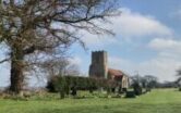 A photograph of a country church, St Peters Corpusty, Norfolk. Set in a green churchyard with blue sky and trees.